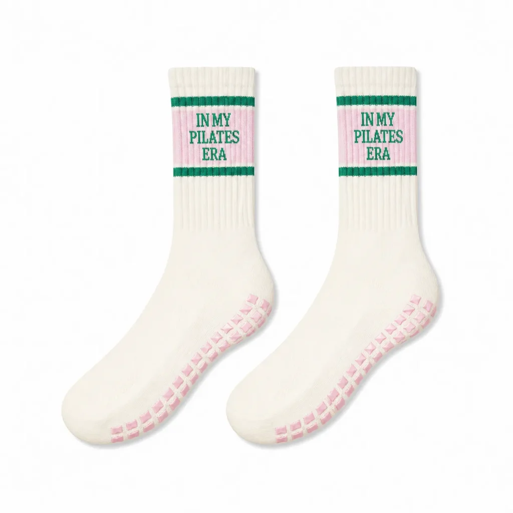 Pair of cream Pilates grip socks with pink text panels, green stripes, and pink silicone grips on a white background.