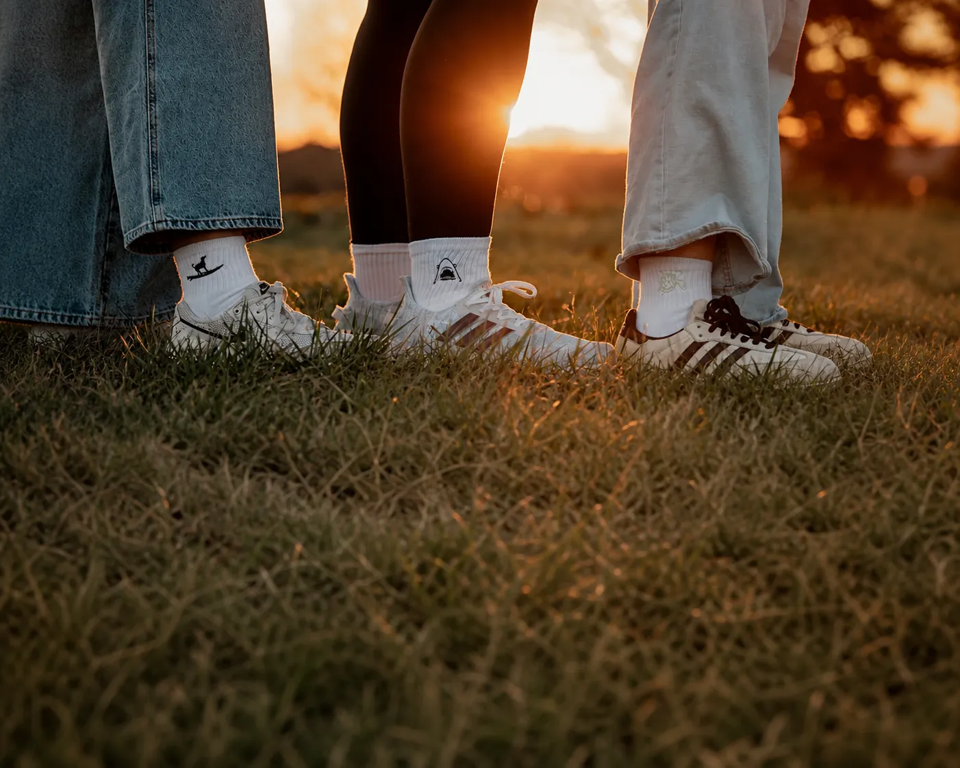 Three people wearing white embroidered logo socks and sneakers on a grassy field at sunset.
