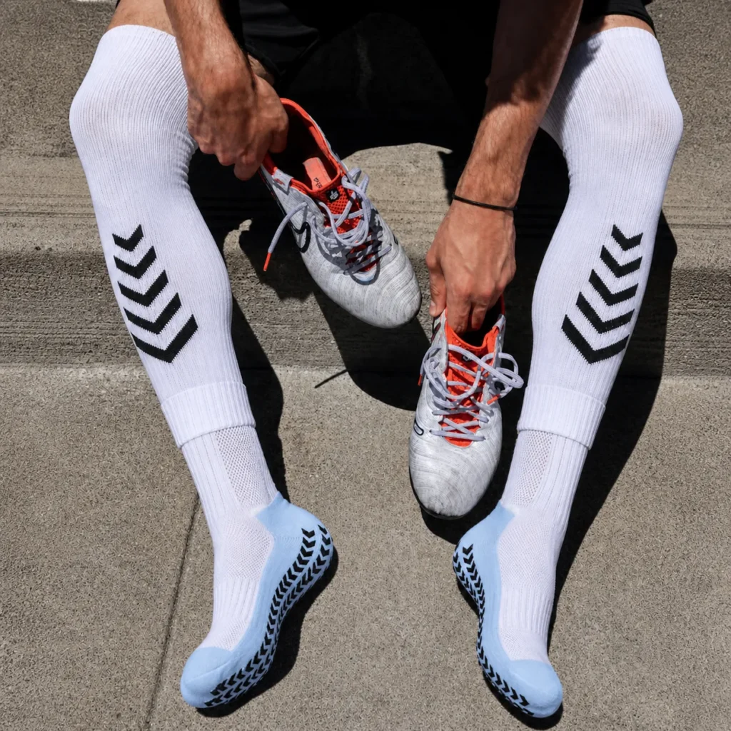 Athlete putting on white soccer grip socks with light blue foot panels and black anti-slip pads.